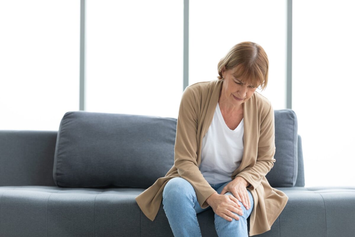 Äldre kvinna sitter på en soffa och håller smärtsamt om sitt knä, ett vanligt tecken på artros och ledvärk.