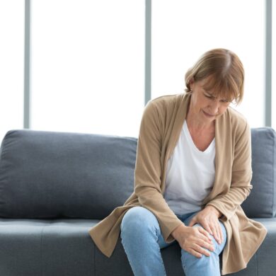 Äldre kvinna sitter på en soffa och håller smärtsamt om sitt knä, ett vanligt tecken på artros och ledvärk.