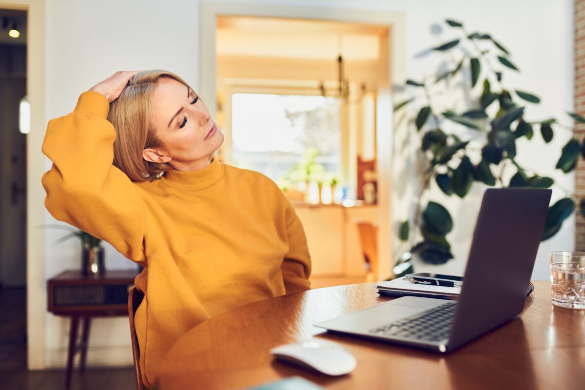 Medelålders kvinna sitter hemma vid datorn, håller sig om nacken och stretchar för att lindra spänningar och förbättra hållningen.