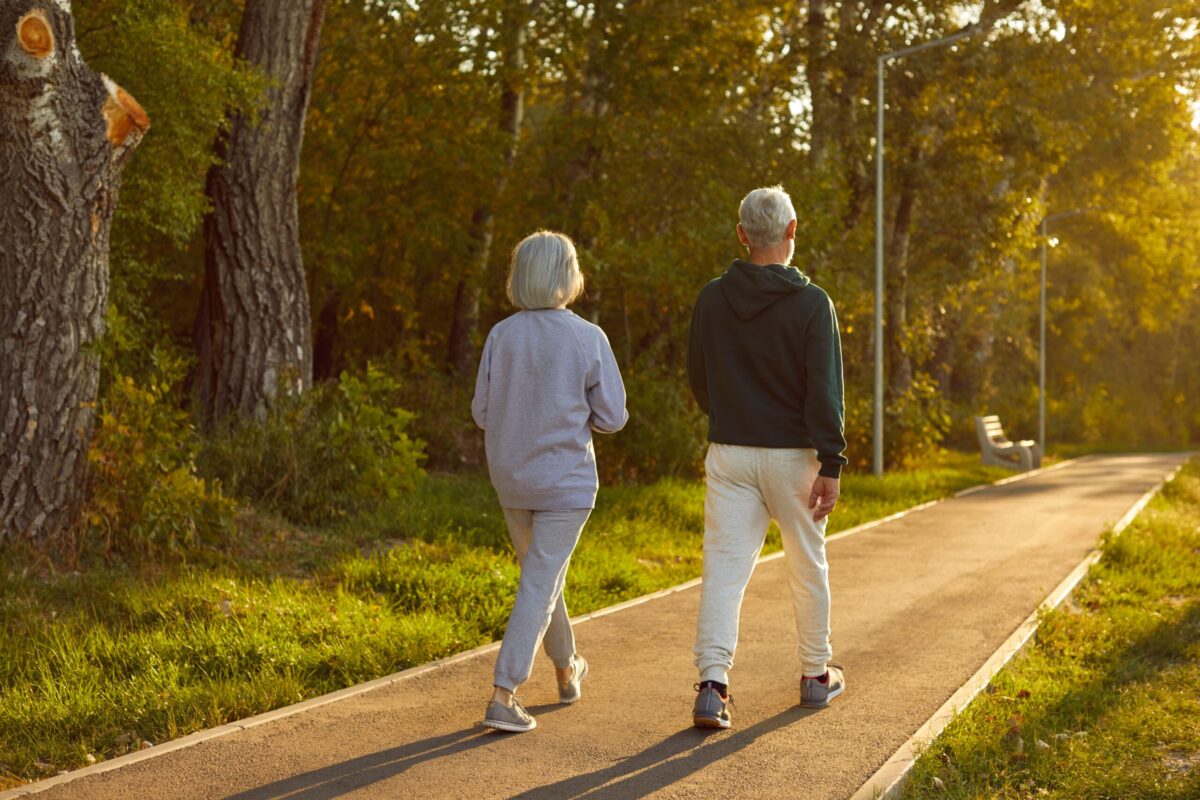 Äldre par, en man och kvinna, promenerar genom skogen med upprätt hållning, sett bakifrån. Deras kroppshållning signalerar ett starkt och hälsosamt skelett.